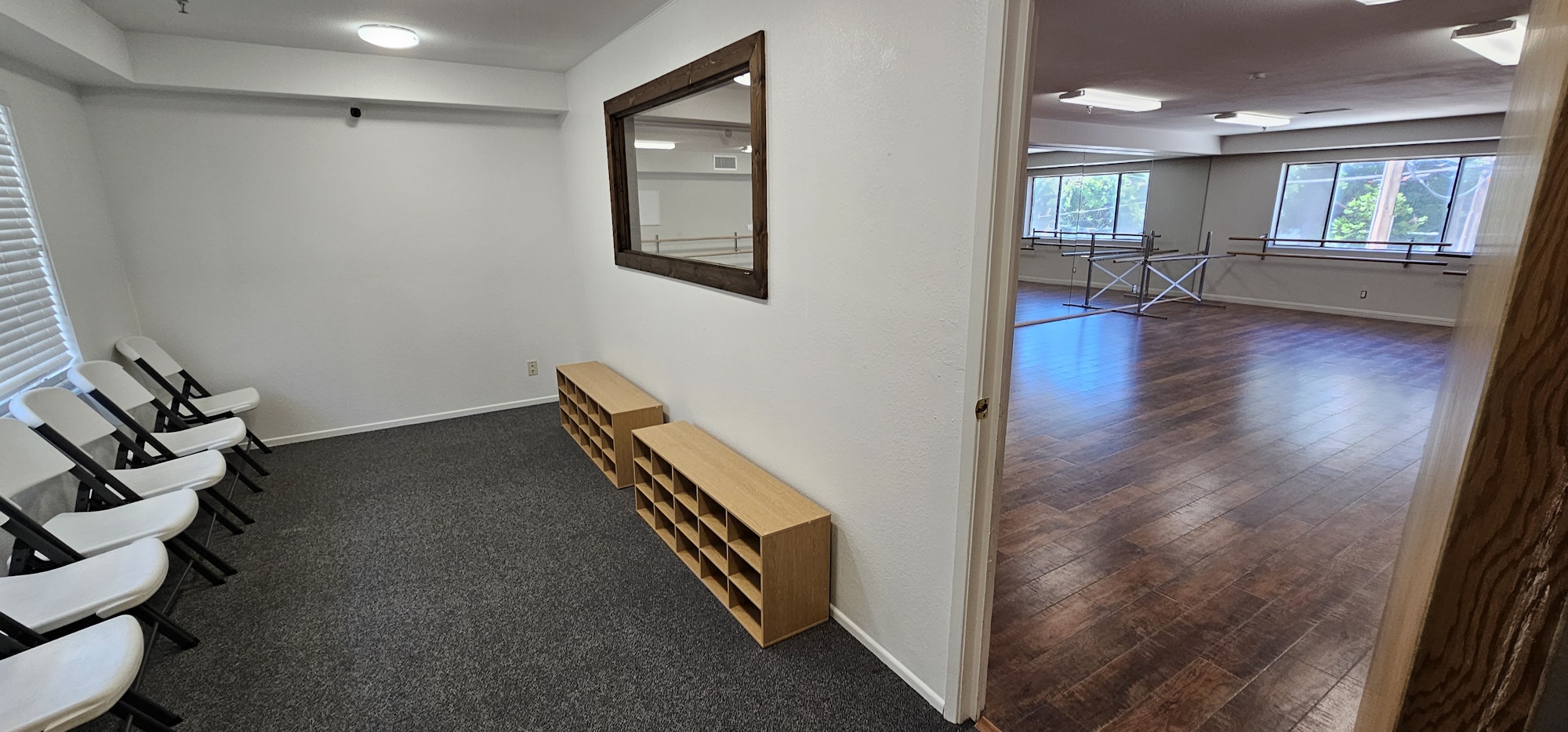 A waiting area with chairs and shelves, leading into a dance studio with wooden floors and mirrors.