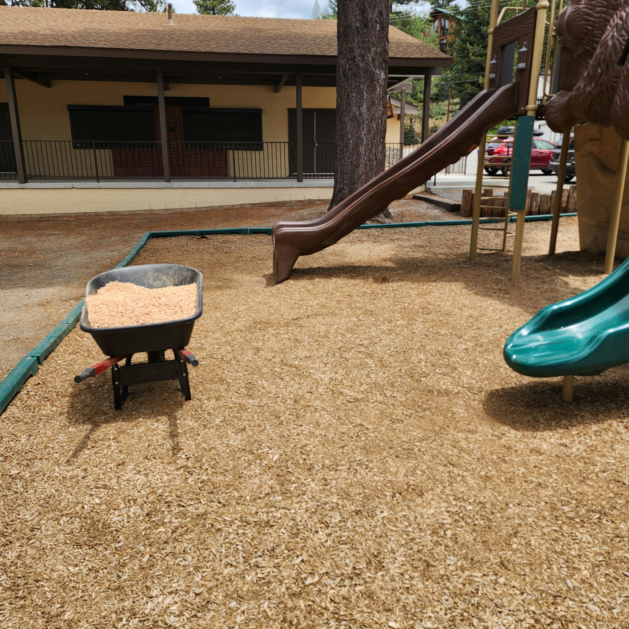 A playground with a wooden and a green slide, a wheelbarrow filled with wood chips, and a building in the background.