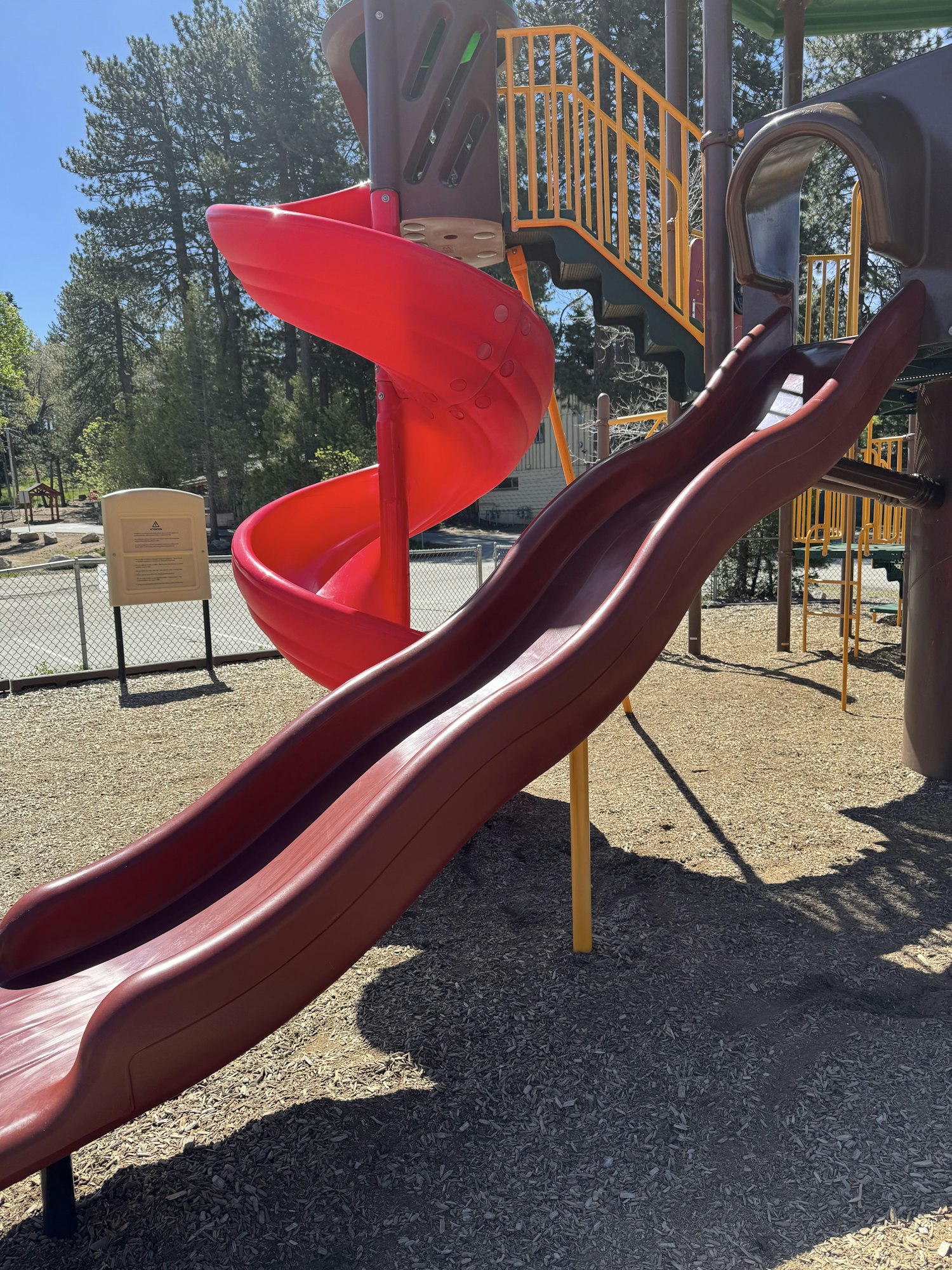 A brightly colored red slide on a playground, with stairs and a gravel surface underneath. Sunlight and trees in the background.