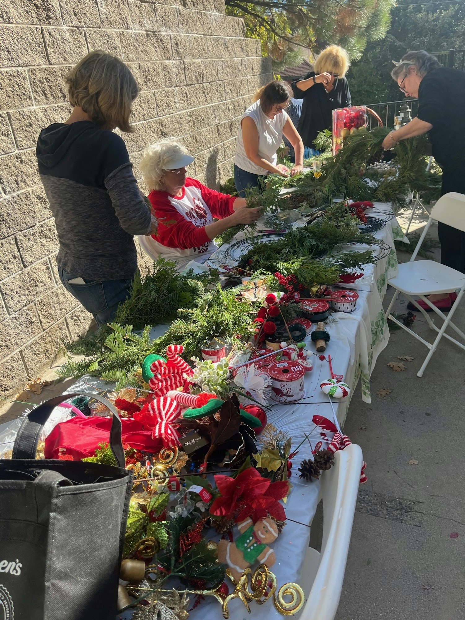 A group of people crafting holiday wreaths outdoors, surrounded by greenery and festive decorations on a table.
