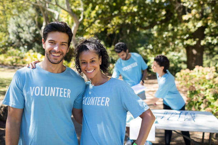 A group of volunteers wearing blue shirts smiles in a park, with others engaged in activities in the background.