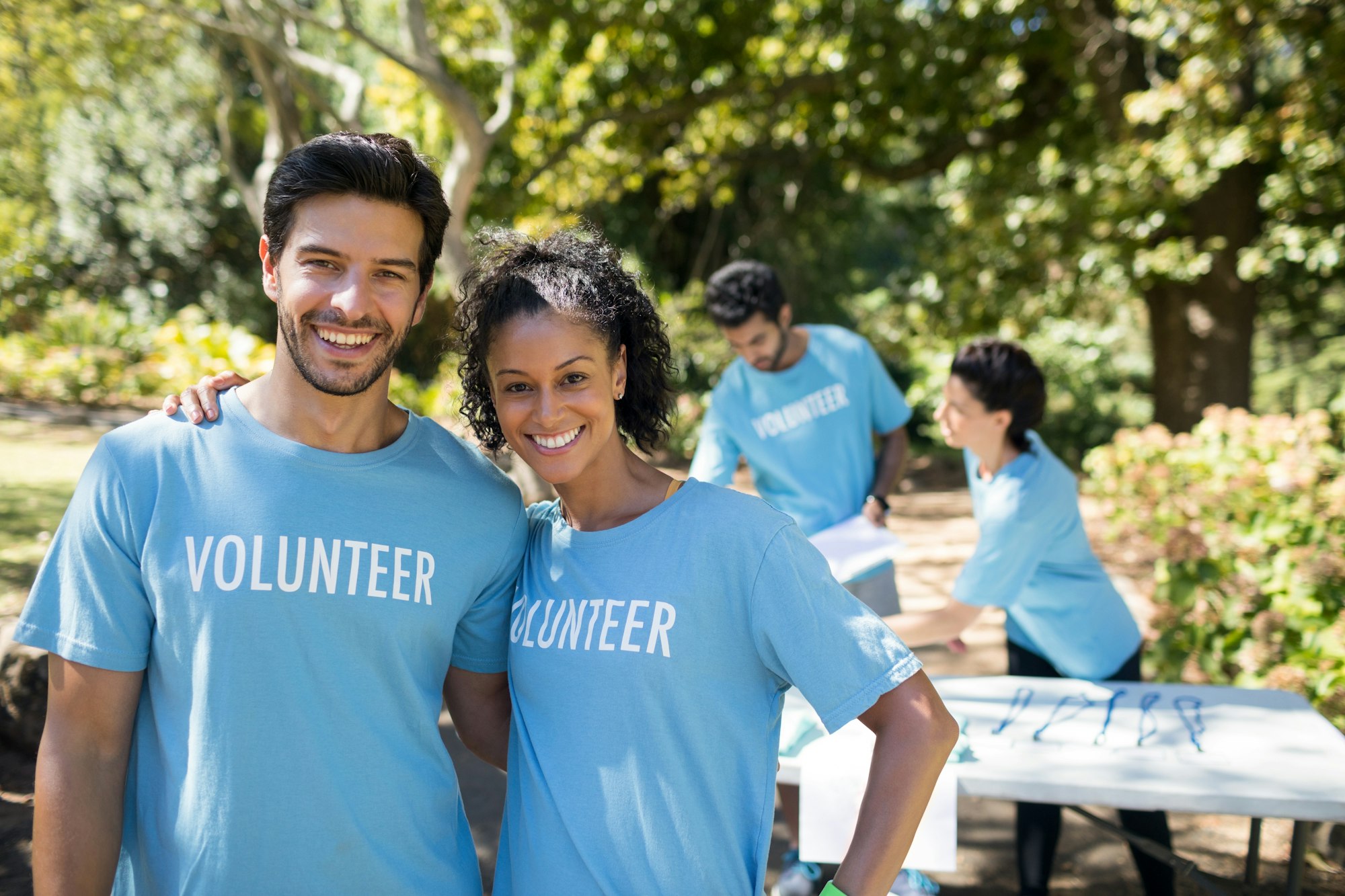 A group of volunteers wearing blue shirts smiles in a park, with others engaged in activities in the background.