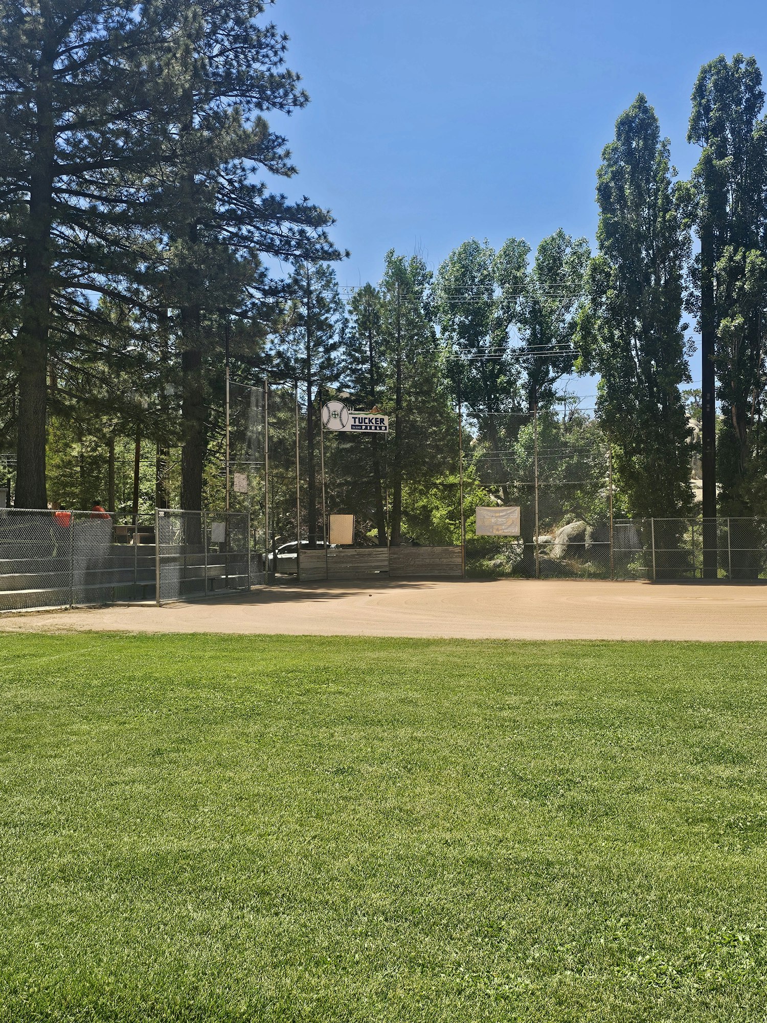 The image shows a baseball field surrounded by trees and a clear blue sky. The grassy area is well maintained.