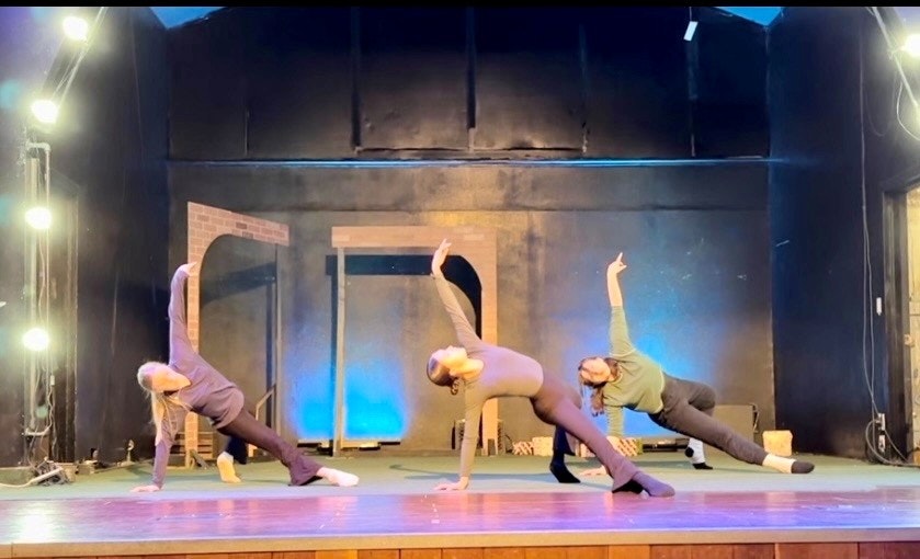 Three people are performing a dance or exercise on a stage, each in a similar pose with one arm extended upward.