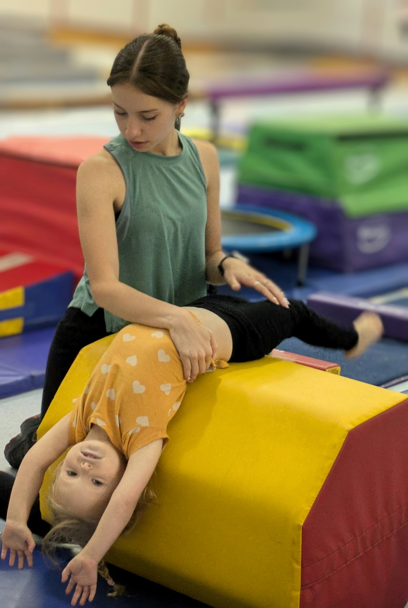 An adult helps a child stretch upside down on a yellow gymnastics pad in a gym.