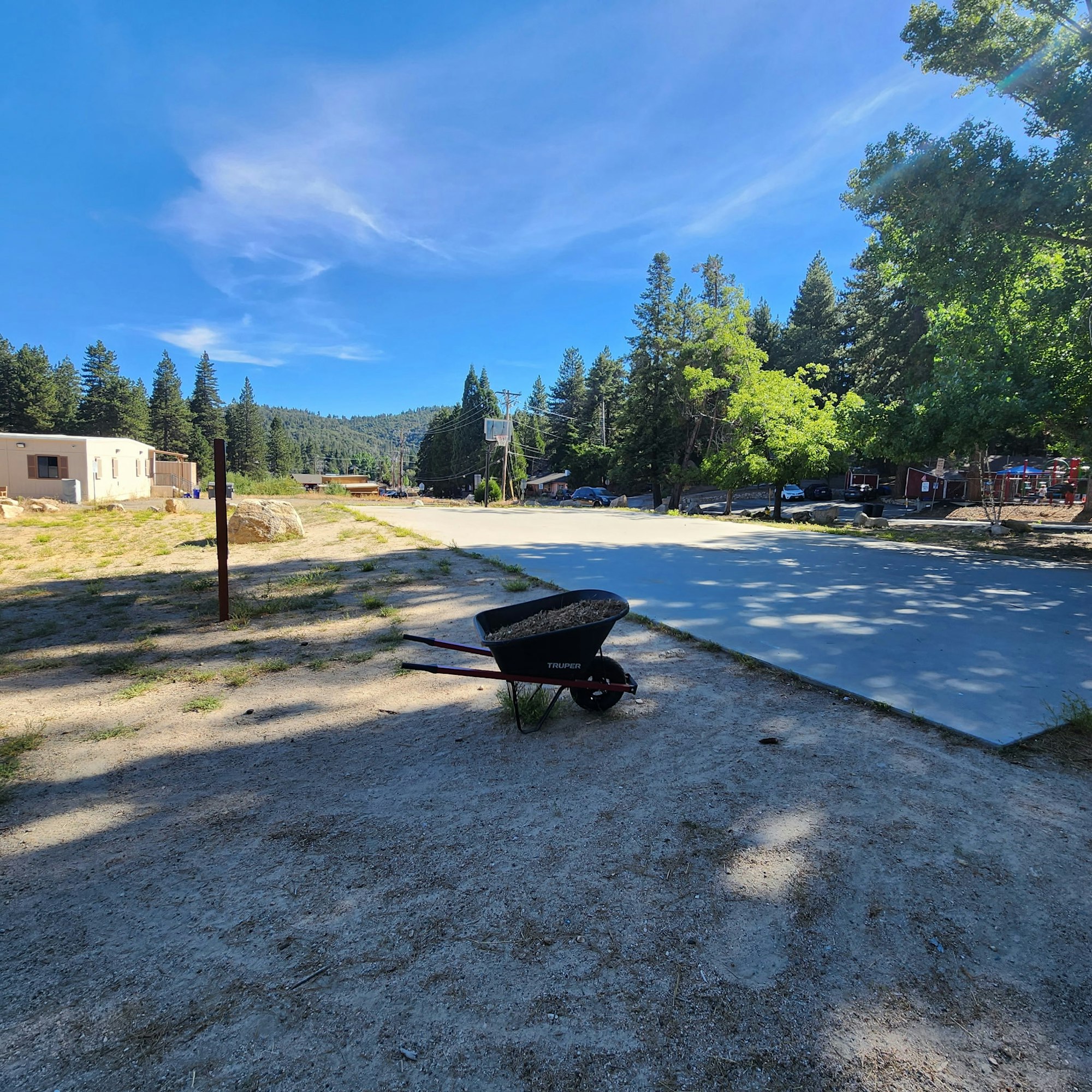 A wheelbarrow sits on a dirt path beside a quiet road, surrounded by trees and distant buildings under a bright blue sky.