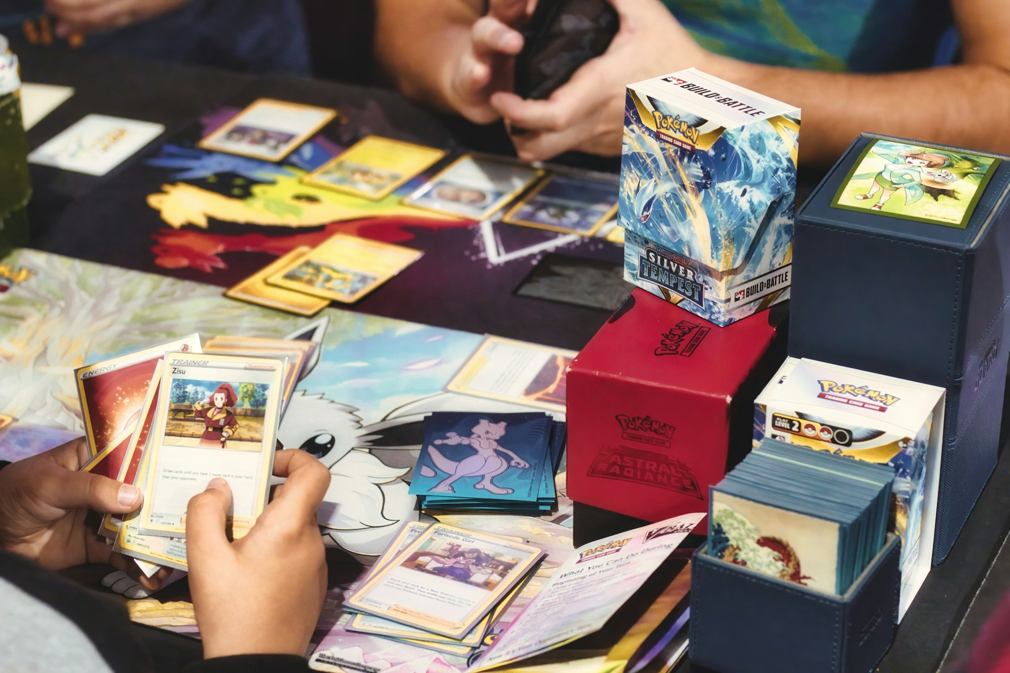 People playing Pokémon trading card game with various cards and boxes on the table.