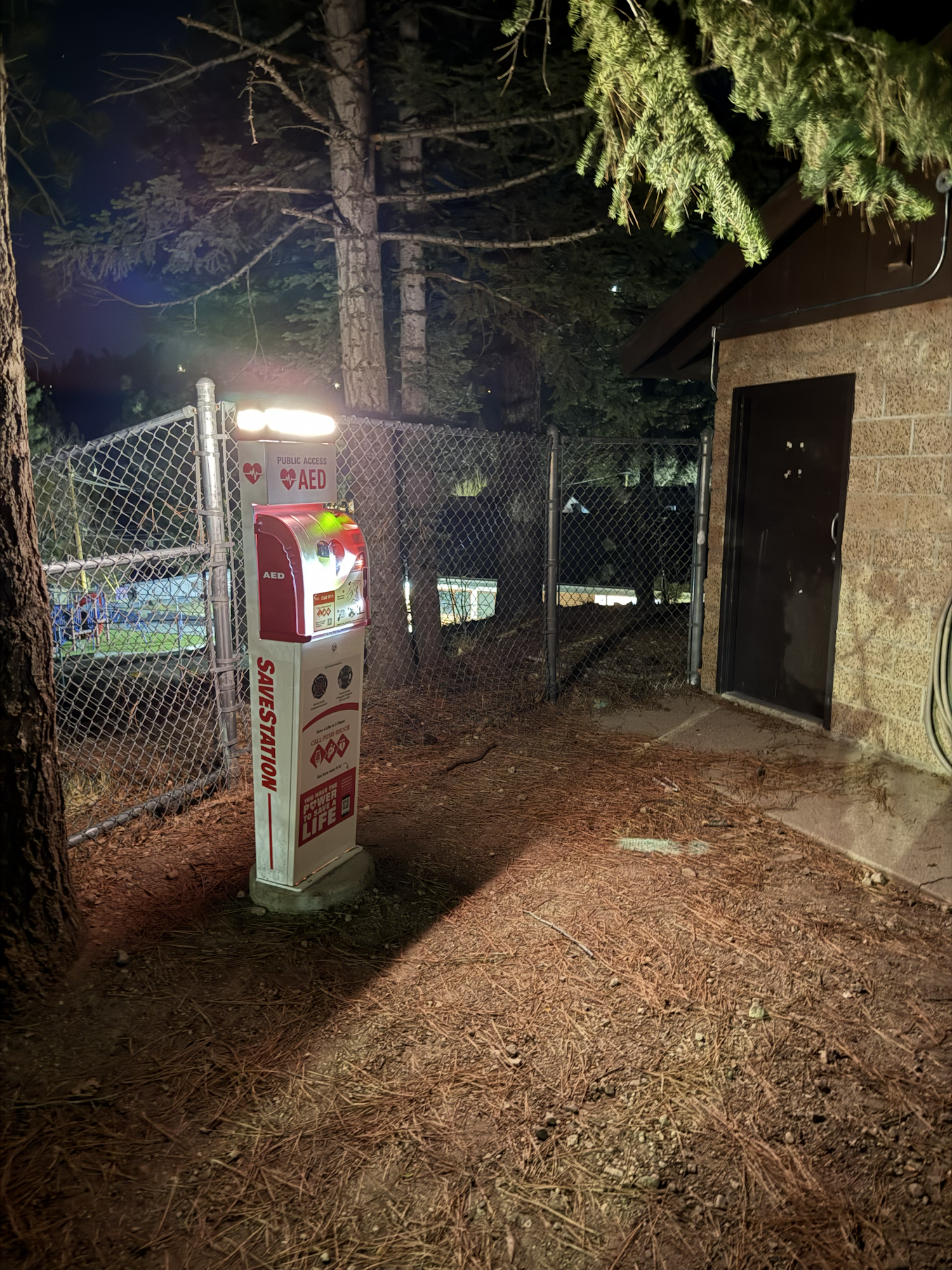 The image shows an AED (Automated External Defibrillator) station illuminated at night, near a fence and surrounded by trees.