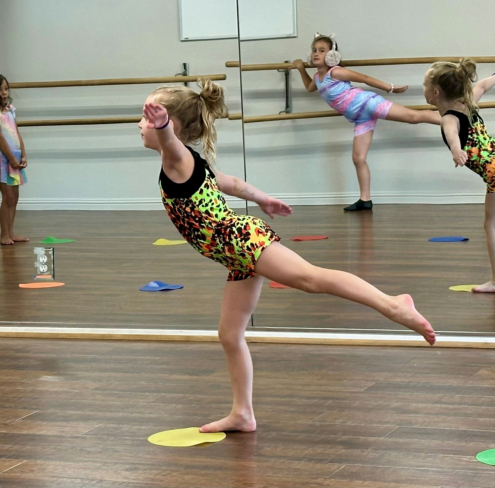 Children practicing ballet poses in a dance studio with mirrors and colorful floor markers.