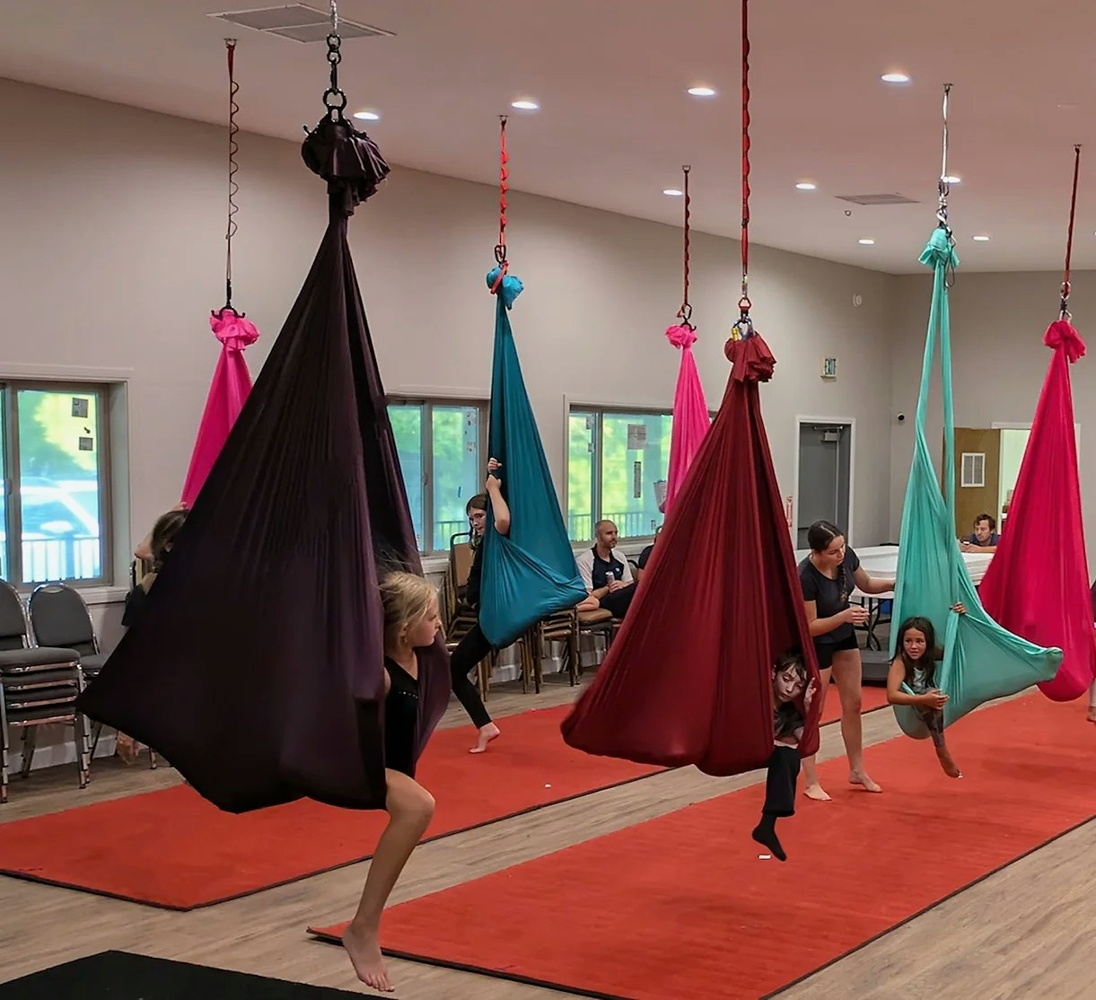 People suspended in colorful aerial hammocks in a room with red mats on the floor.