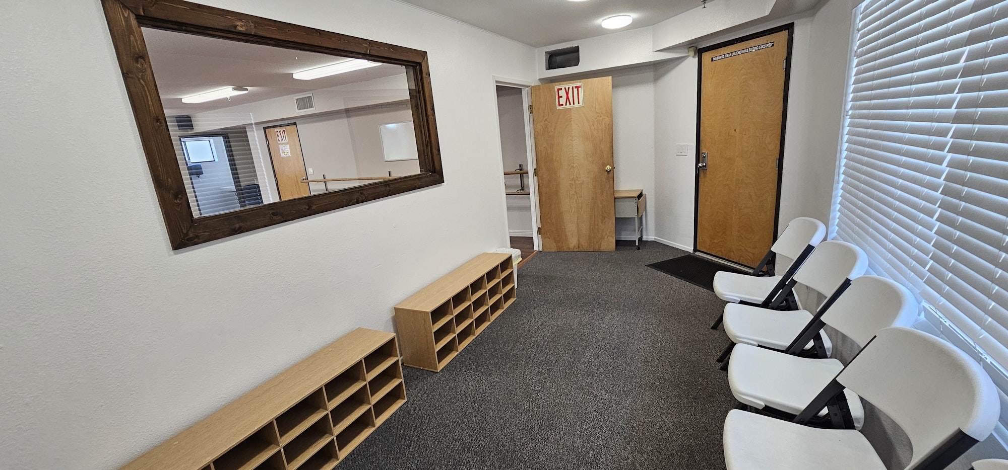A waiting room with white folding chairs, a large wall mirror, shoe racks, and an exit door.