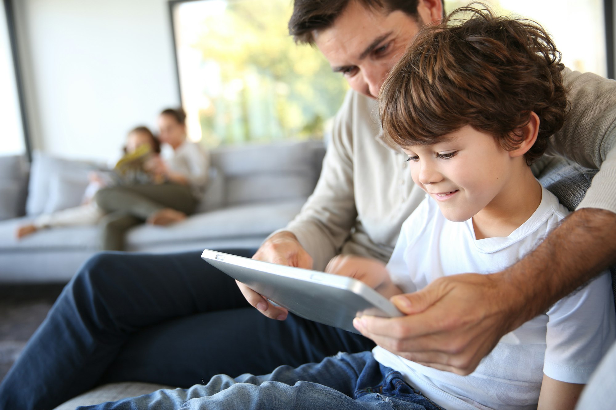 Father and son using a tablet on a couch, while a mother reads with another child in the background.