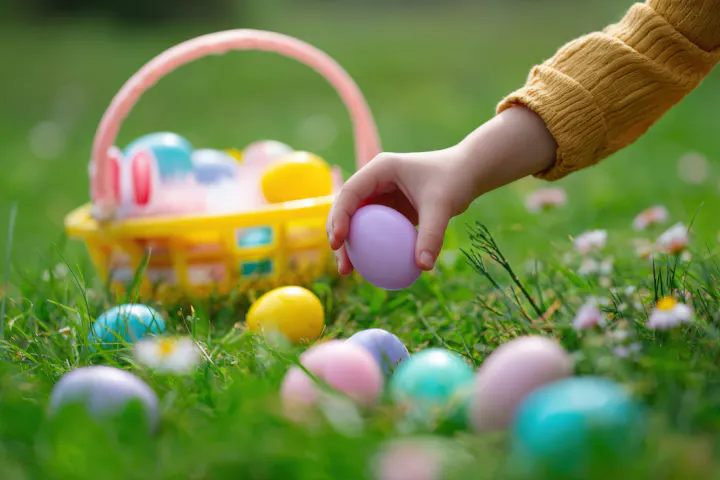 A hand is picking up a purple Easter egg from grass, near a colorful basket filled with eggs.