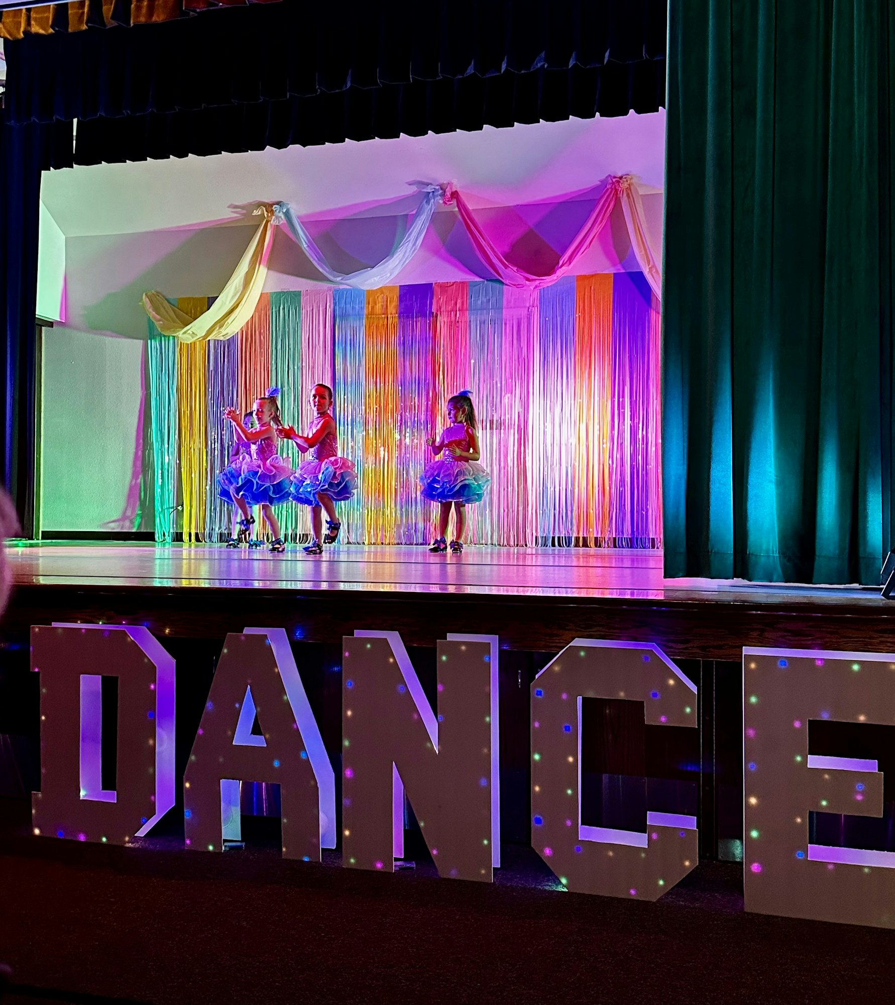 A stage with colorful curtains and dancers in vibrant costumes. Large illuminated "DANCE" letters are in front.
