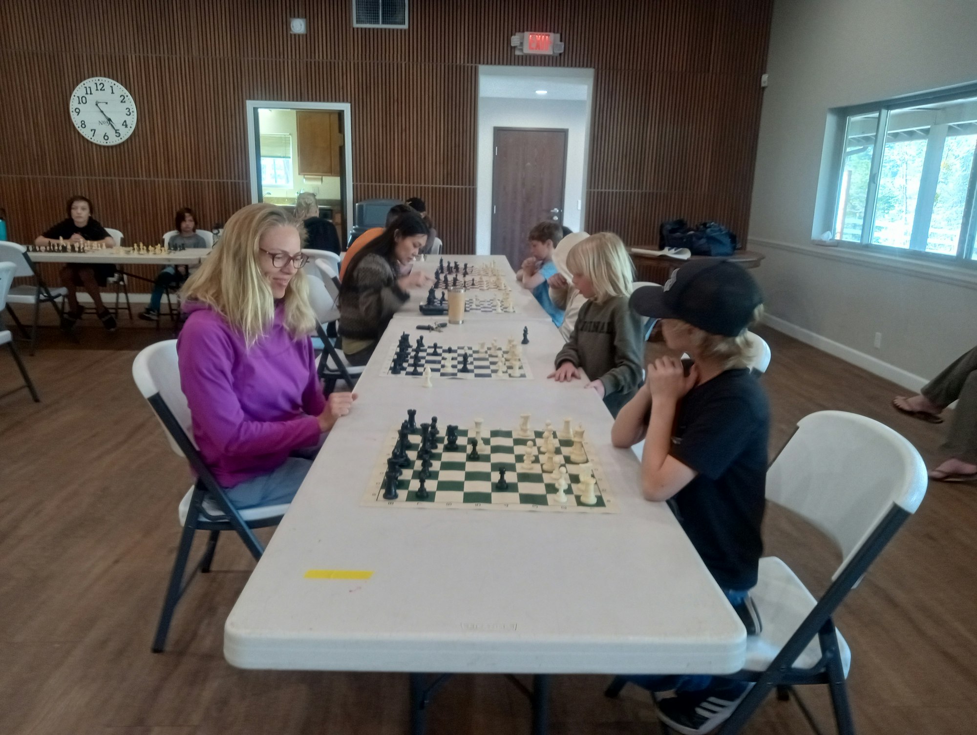 People playing chess at a long table in a room with wooden walls and a clock.
