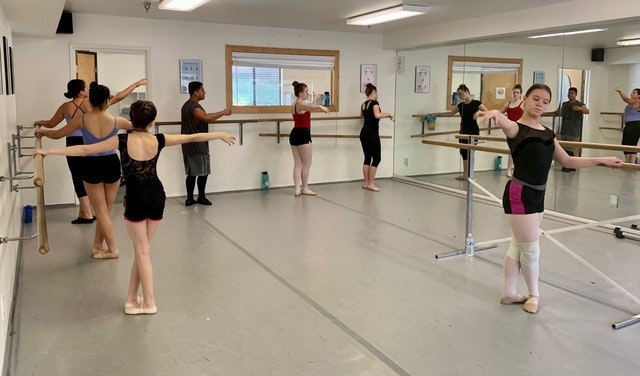 Ballet class with dancers practicing at barres in a mirrored dance studio.