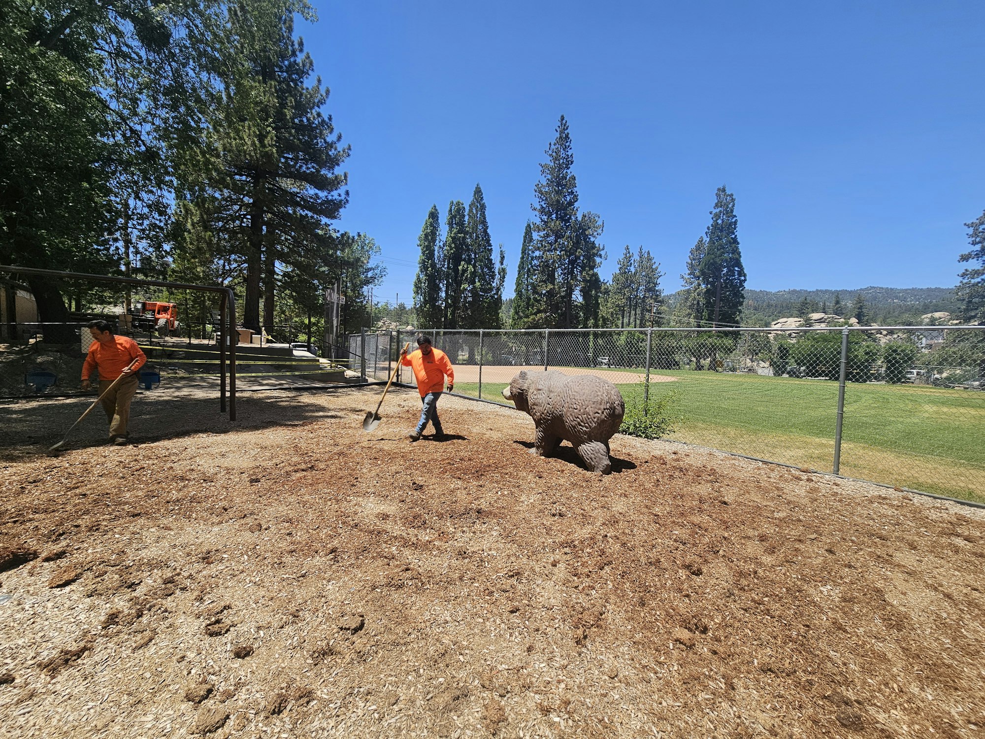 Two people raking soil near a sculpture of a bear in a fenced area with trees in the background.