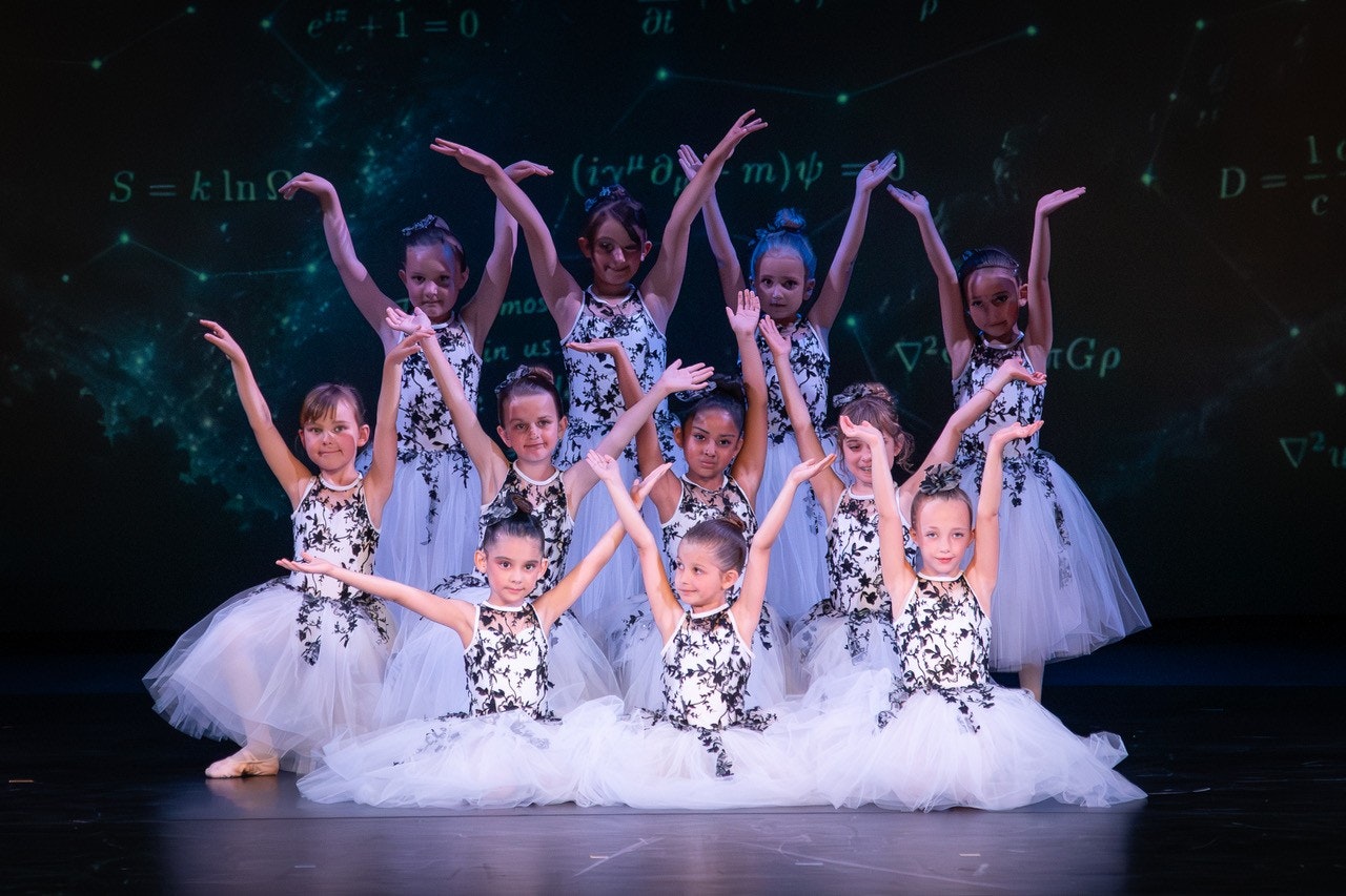 A group of young girls in elegant, floral-patterned ballet dresses posing on stage with graceful hand movements.