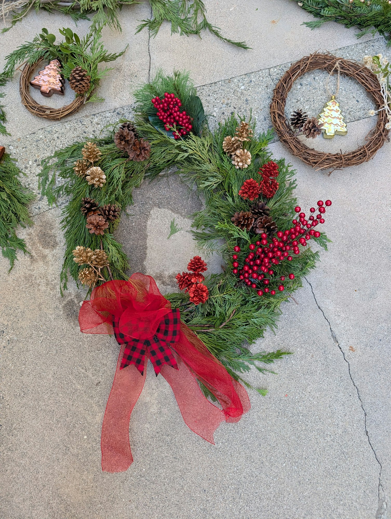 Festive wreaths with pinecones, red berries, and a red bow on a concrete surface.