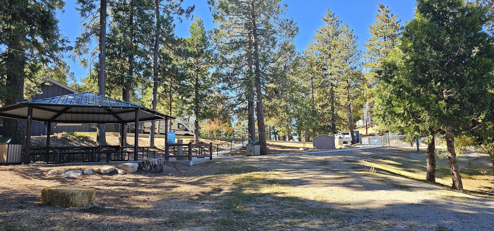 A sunny outdoor area with a gazebo, picnic tables, trees, and a gravel path, suggesting a recreational space.