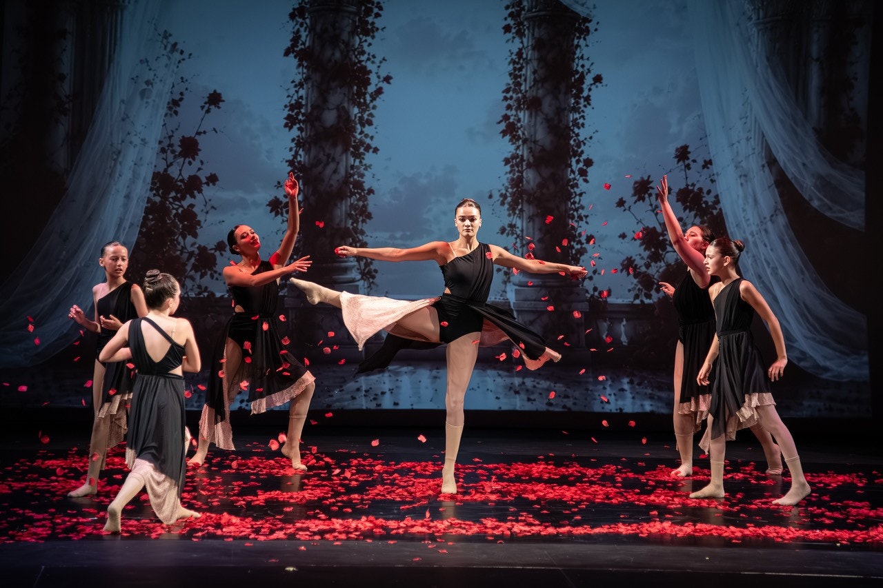 A ballet performance featuring dancers in black dresses on a stage scattered with rose petals, set against a scenic backdrop.