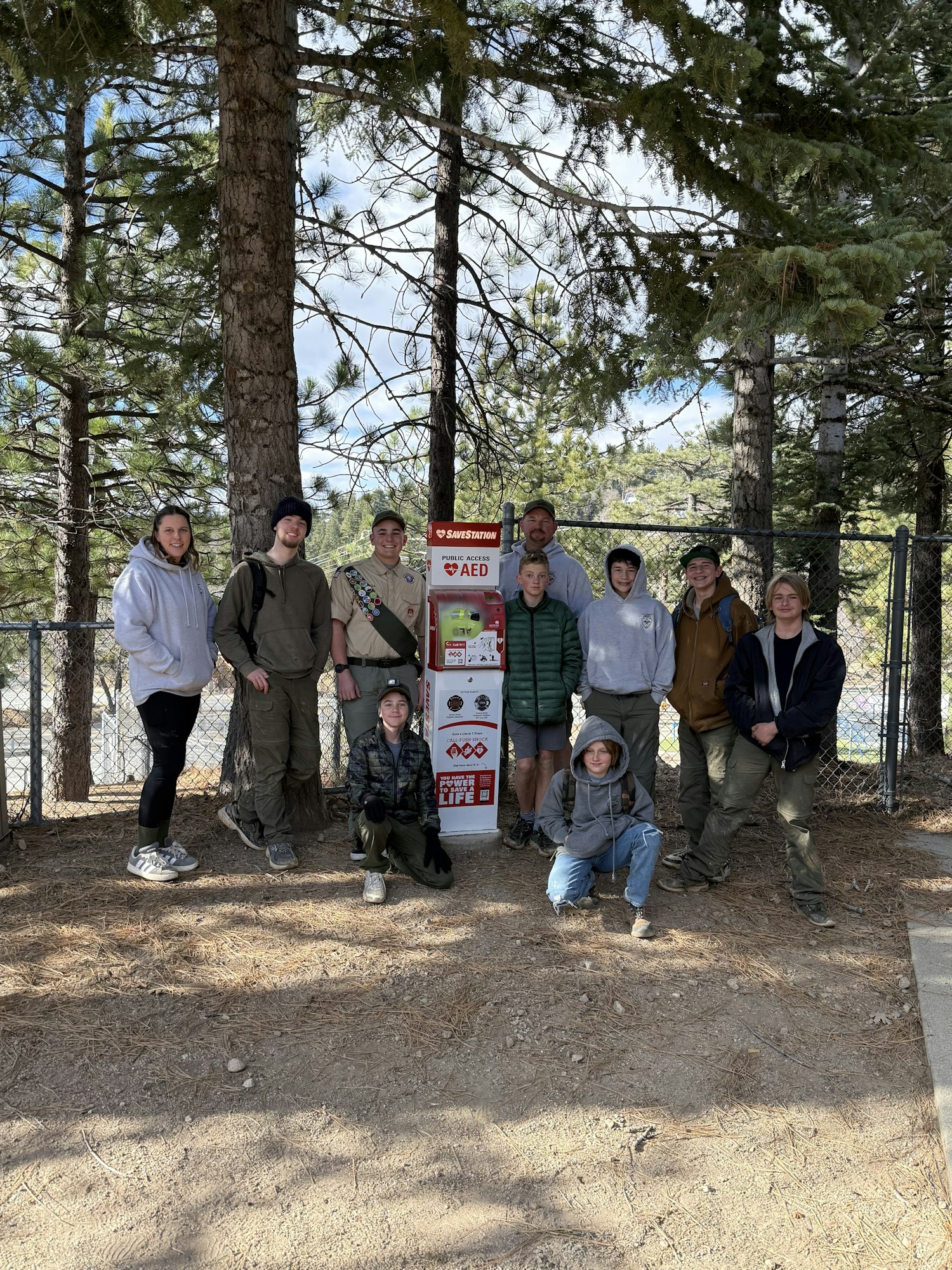 A group of people gathered outdoors near a sign, surrounded by trees, with some sitting and others standing in casual attire.