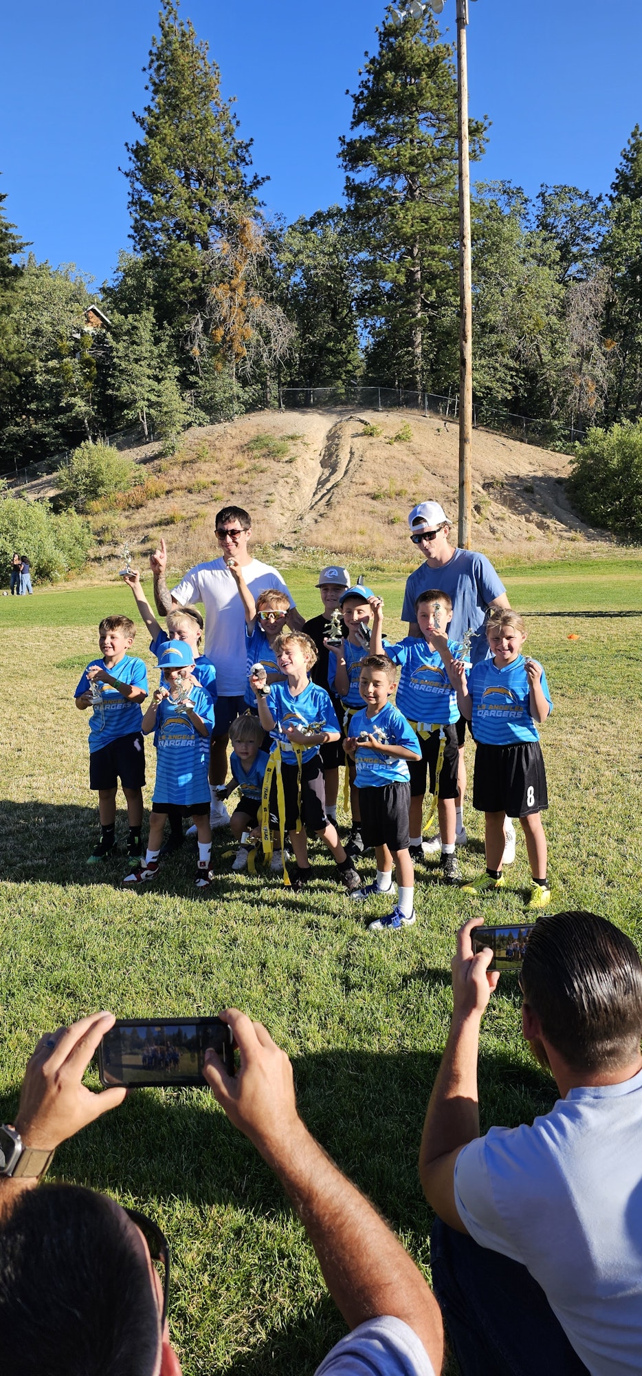 A group of children in blue sports shirts posing for a photo with trophies, celebrating a team accomplishment in a park.