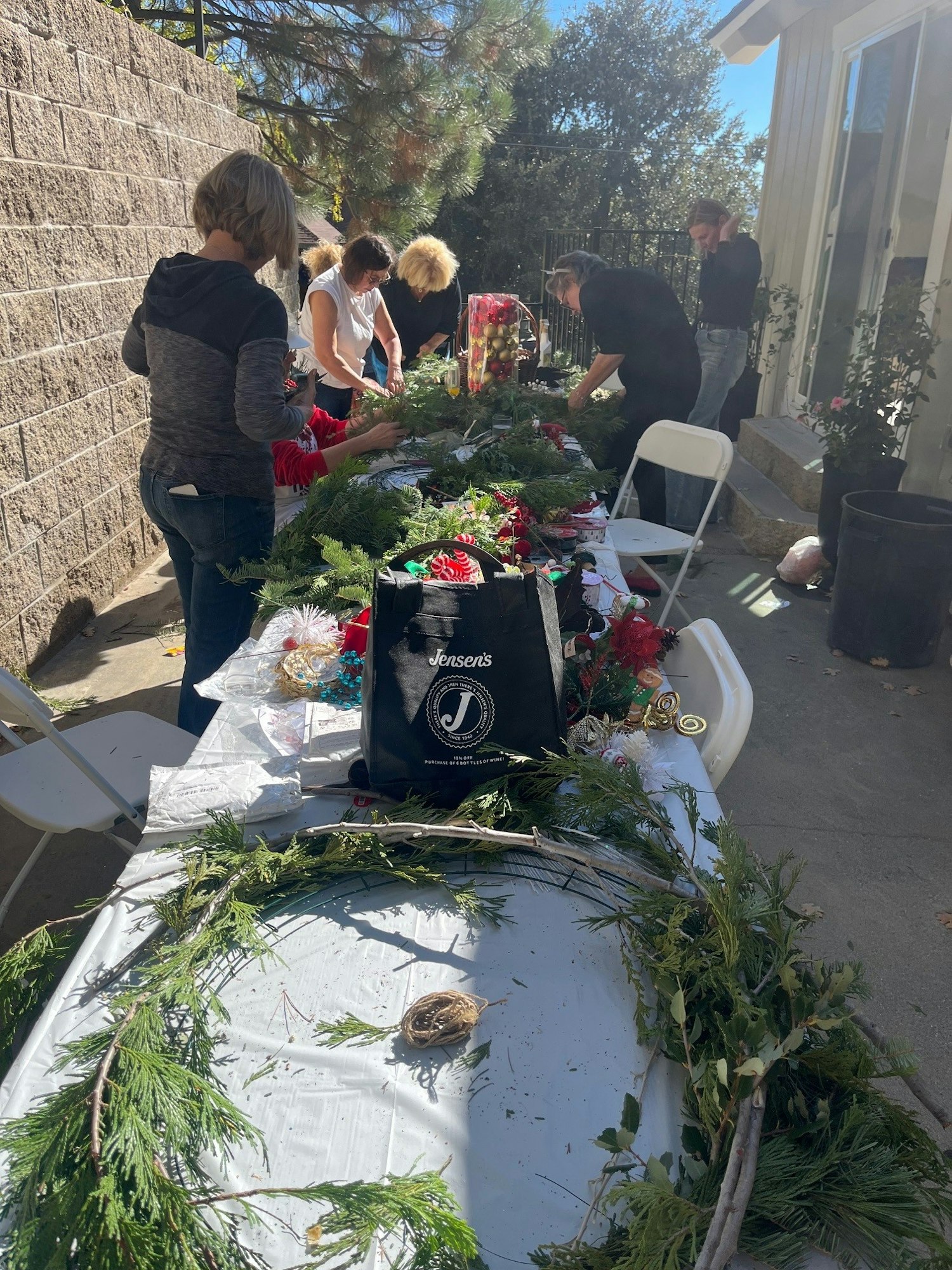 People crafting wreaths at a table outdoors, with greenery and decorative supplies spread around.