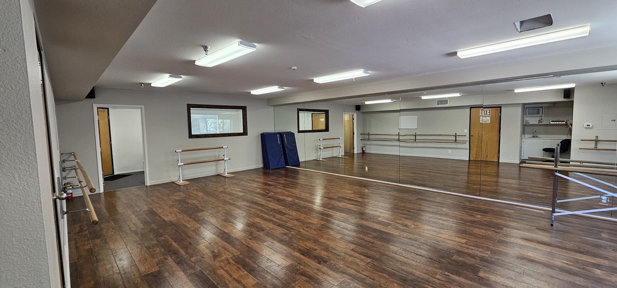 Spacious dance studio with wooden floors, mirrors, ballet barres, and fluorescent lighting.