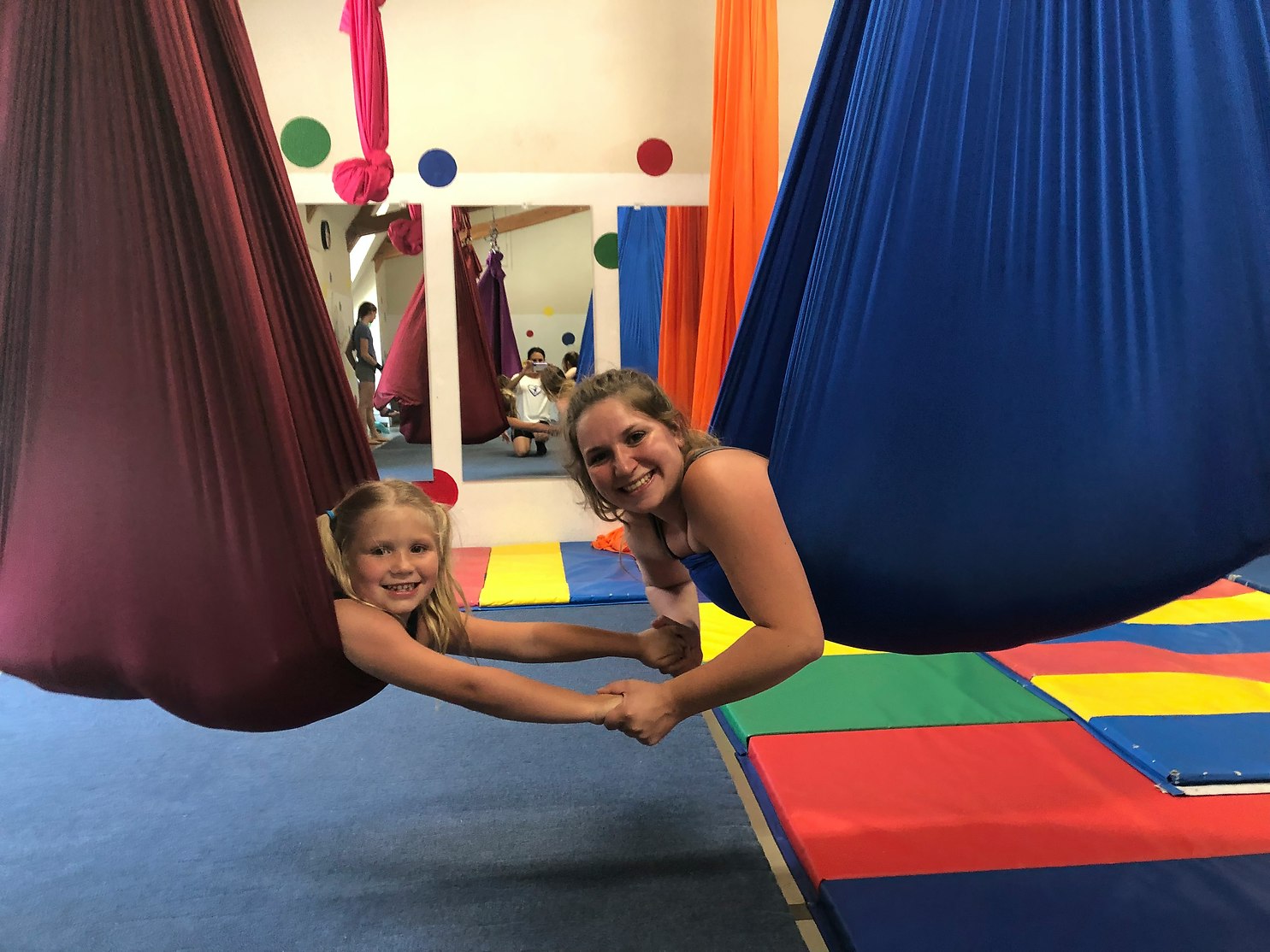 Two people smiling and holding hands in colorful hammock swings indoors.