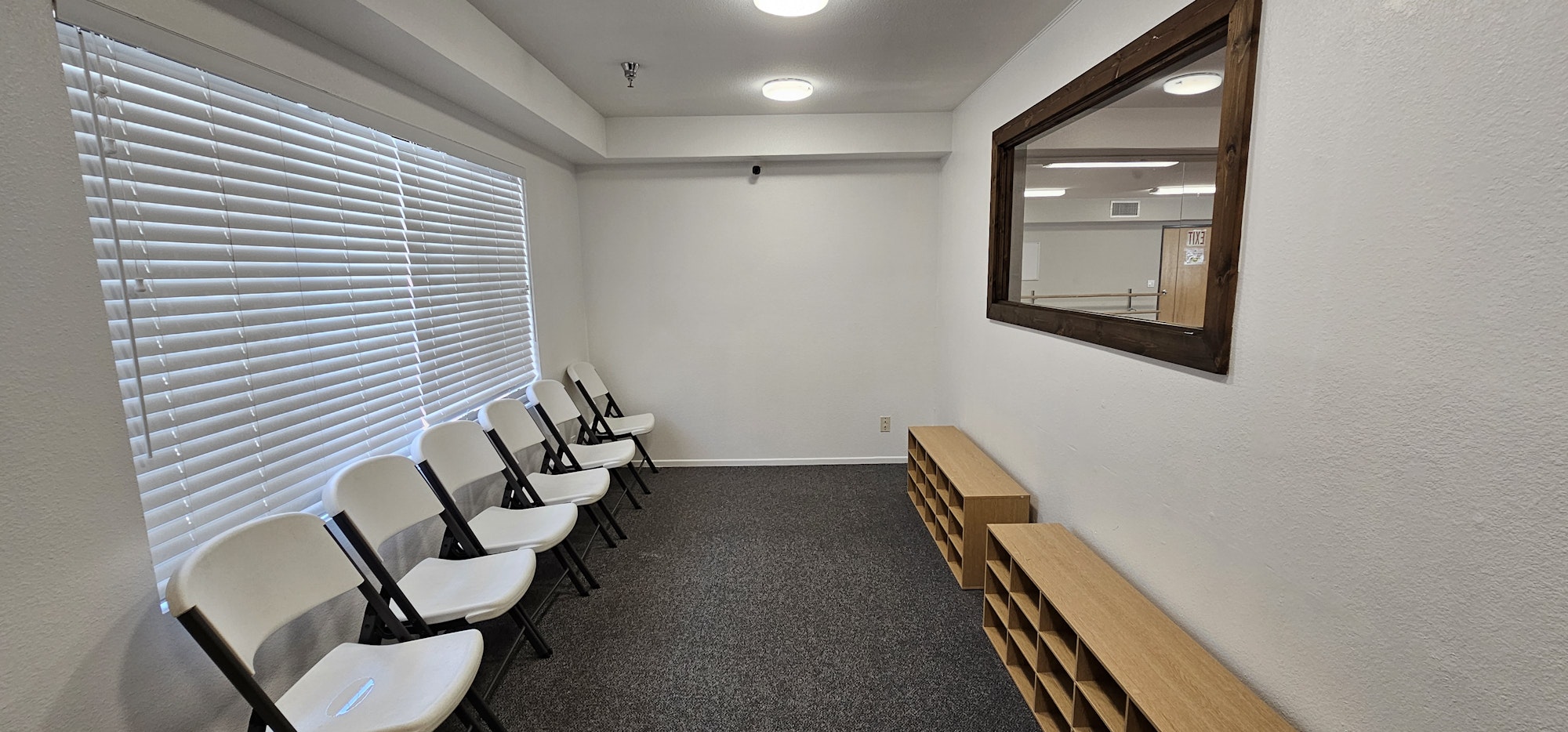 A waiting room with white folding chairs, a large mirror, blinds, and wooden storage units on a carpeted floor.