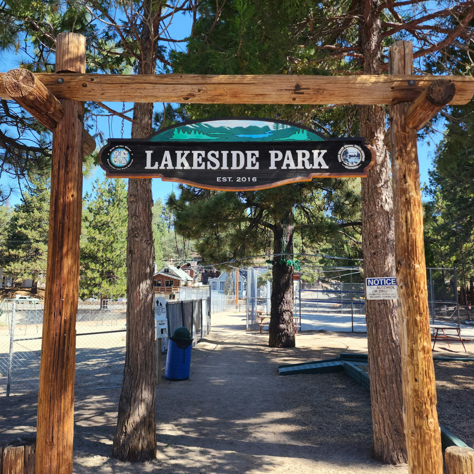 A wooden sign marks the entrance to Lakeside Park, established in 2016, surrounded by trees and a fenced area.