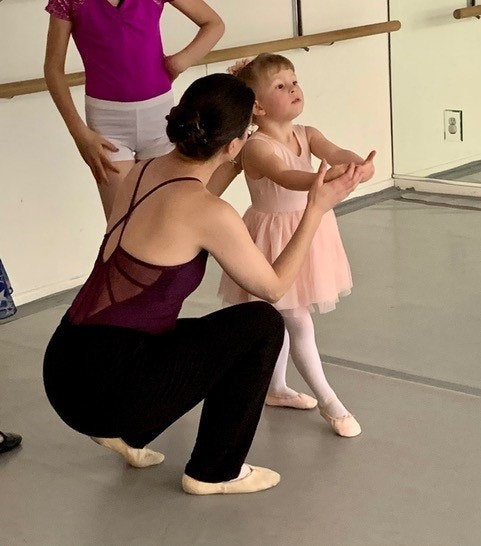 A dance instructor guides a young girl in ballet posture, with another child watching in a dance studio.