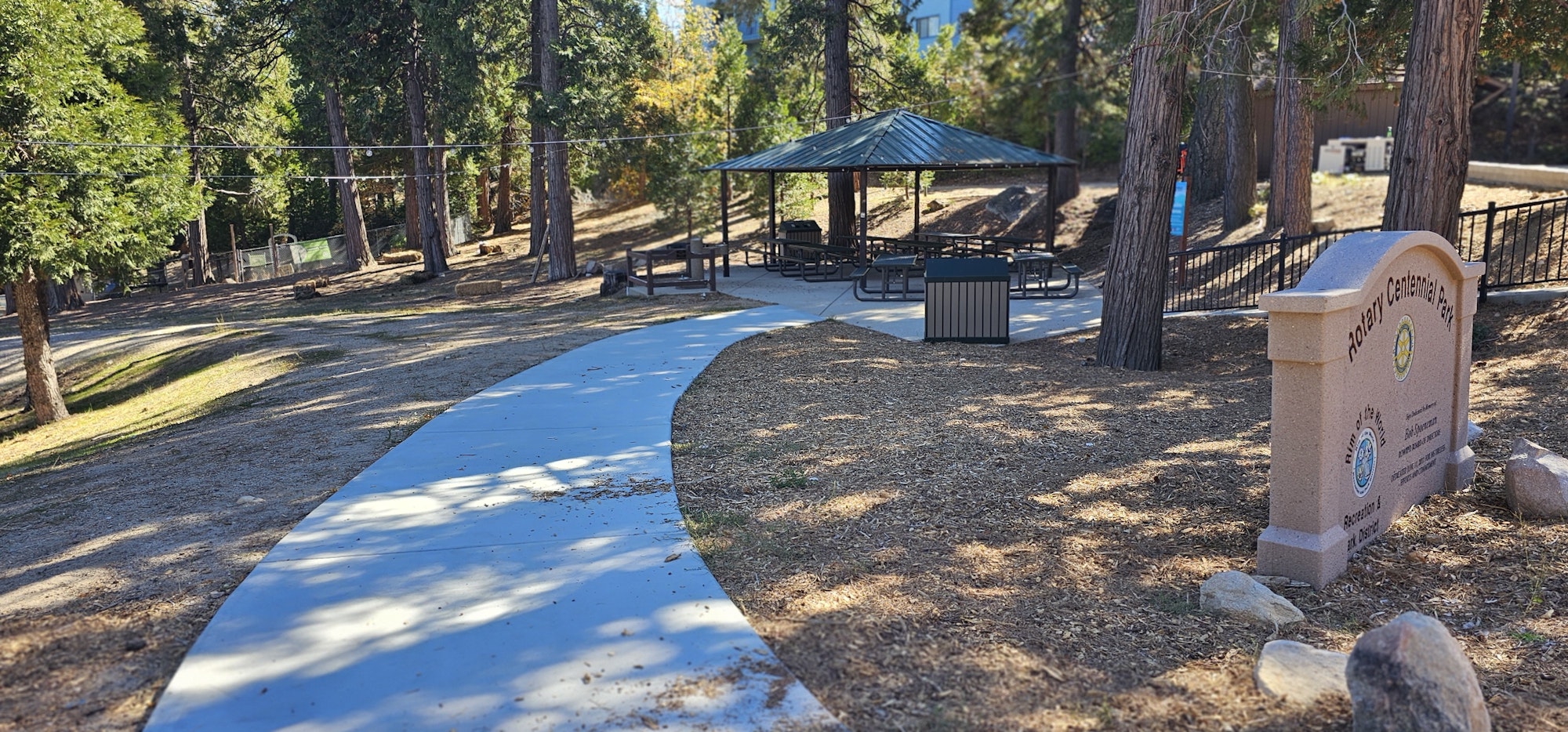 The image shows a park pathway leading to a gazebo with picnic tables, surrounded by trees and a park sign.