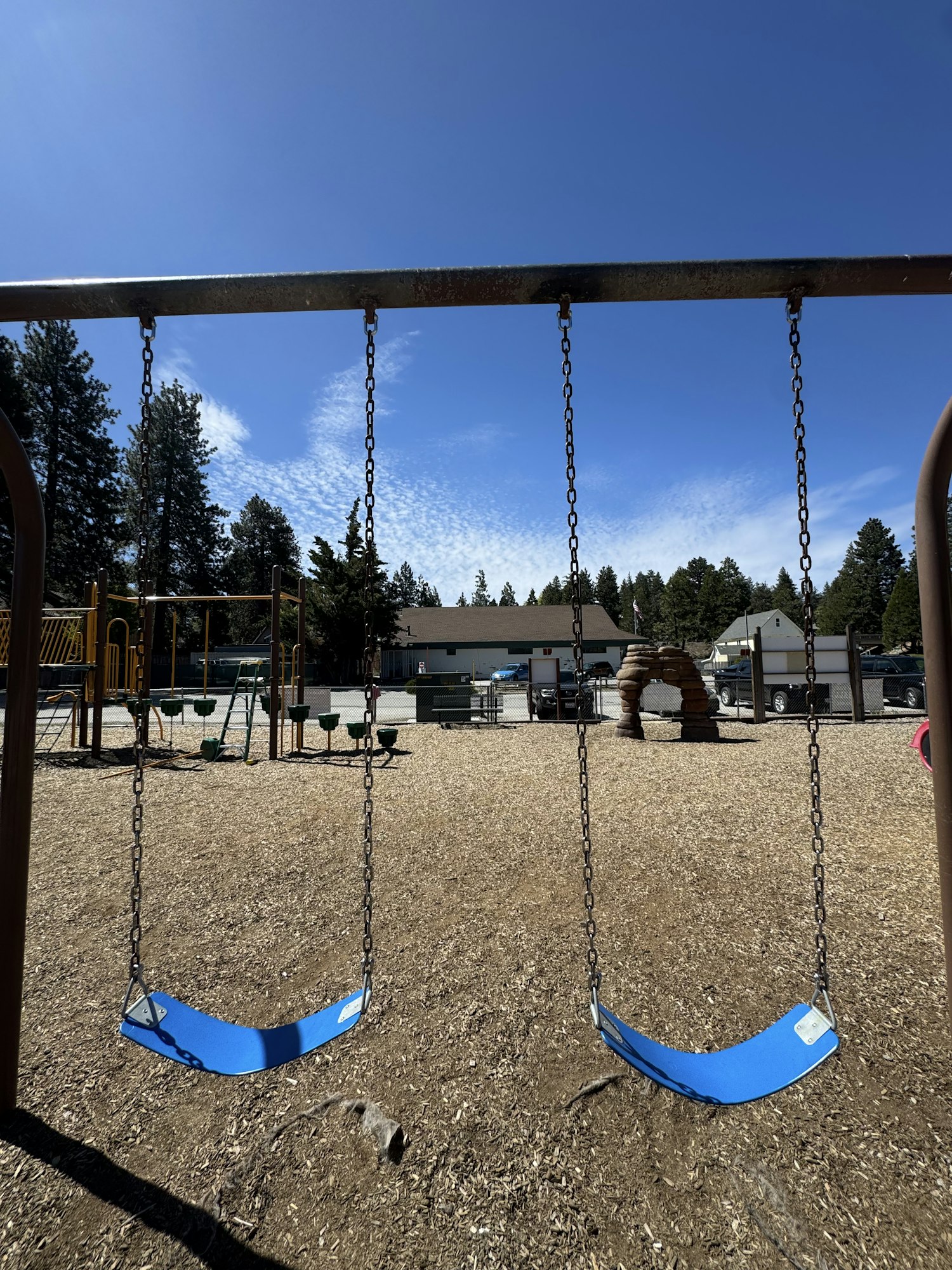 An empty playground featuring blue swings, a clear blue sky, and trees in the background.