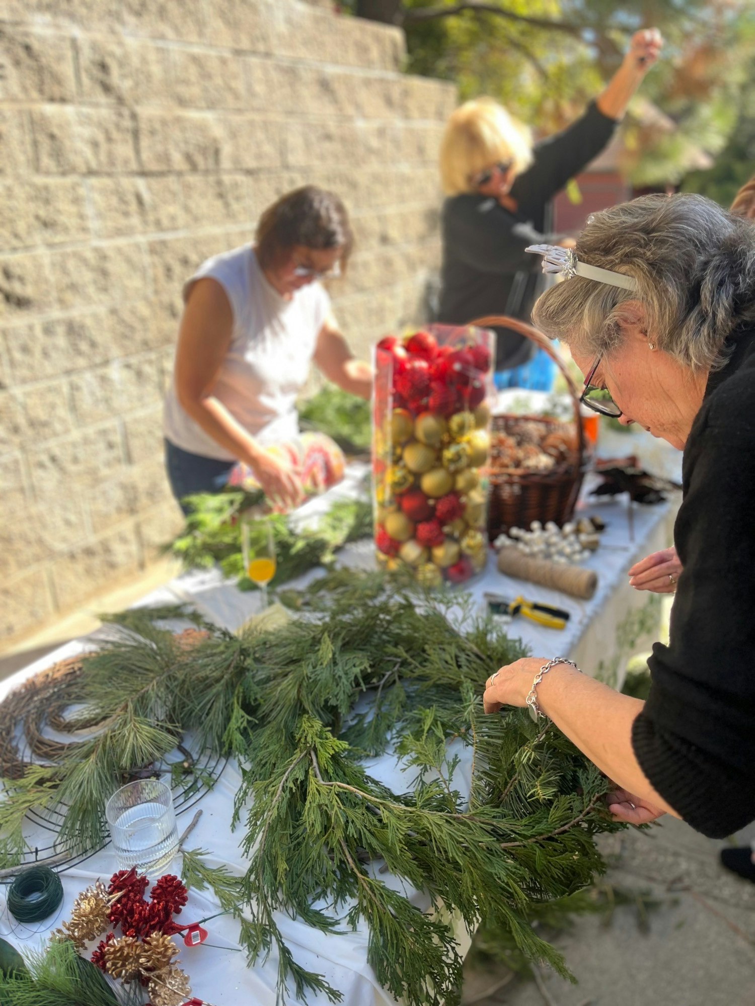 Three people making wreaths with greenery and decorative items on a table outdoors.