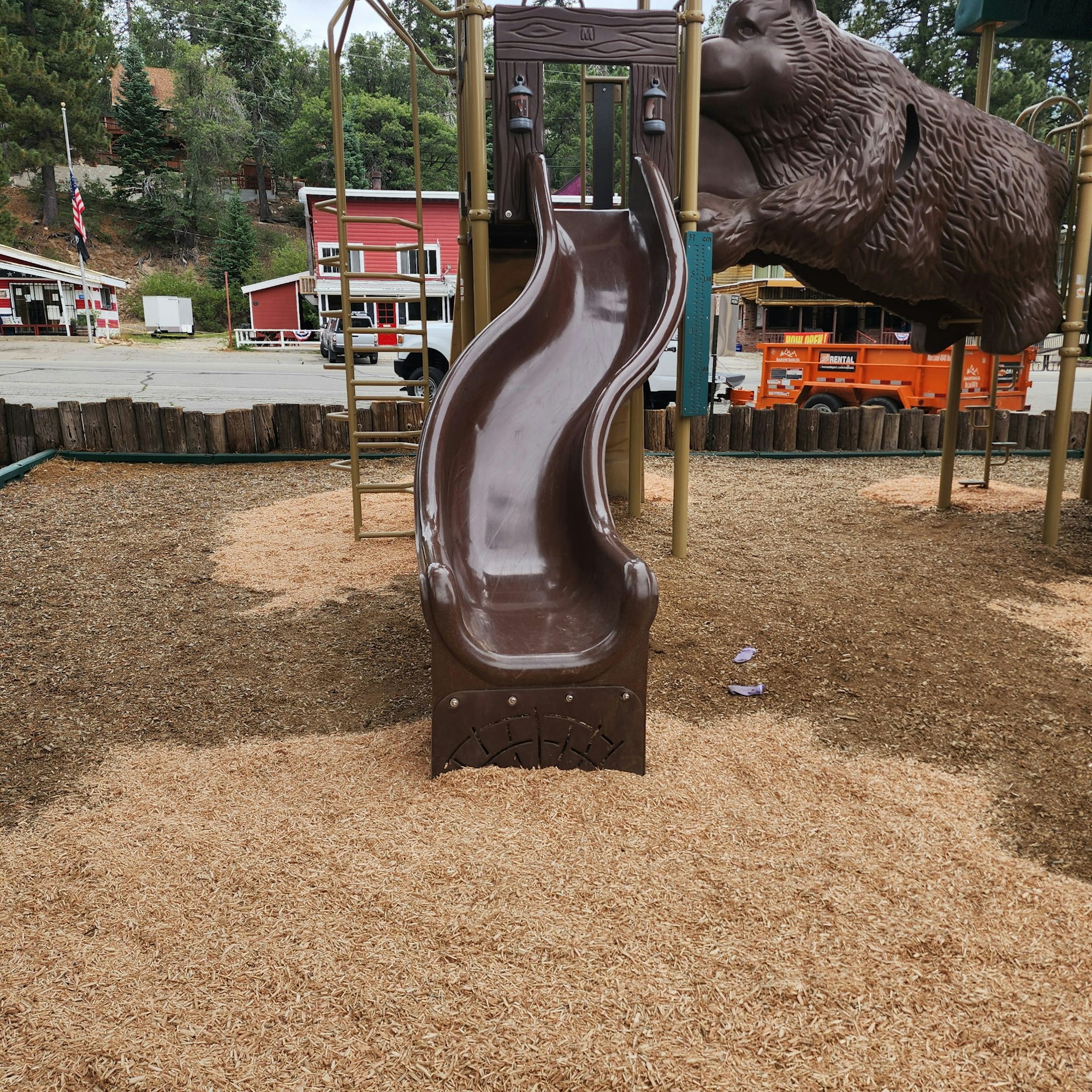 A playground slide with a bear design, surrounded by wood chips, near some buildings and parked vehicles in a forested area.