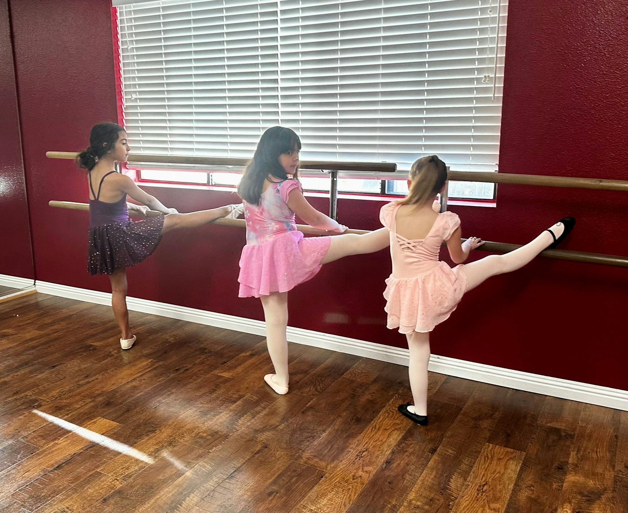 Three children in ballet outfits practice leg stretches on a barre in a dance studio.