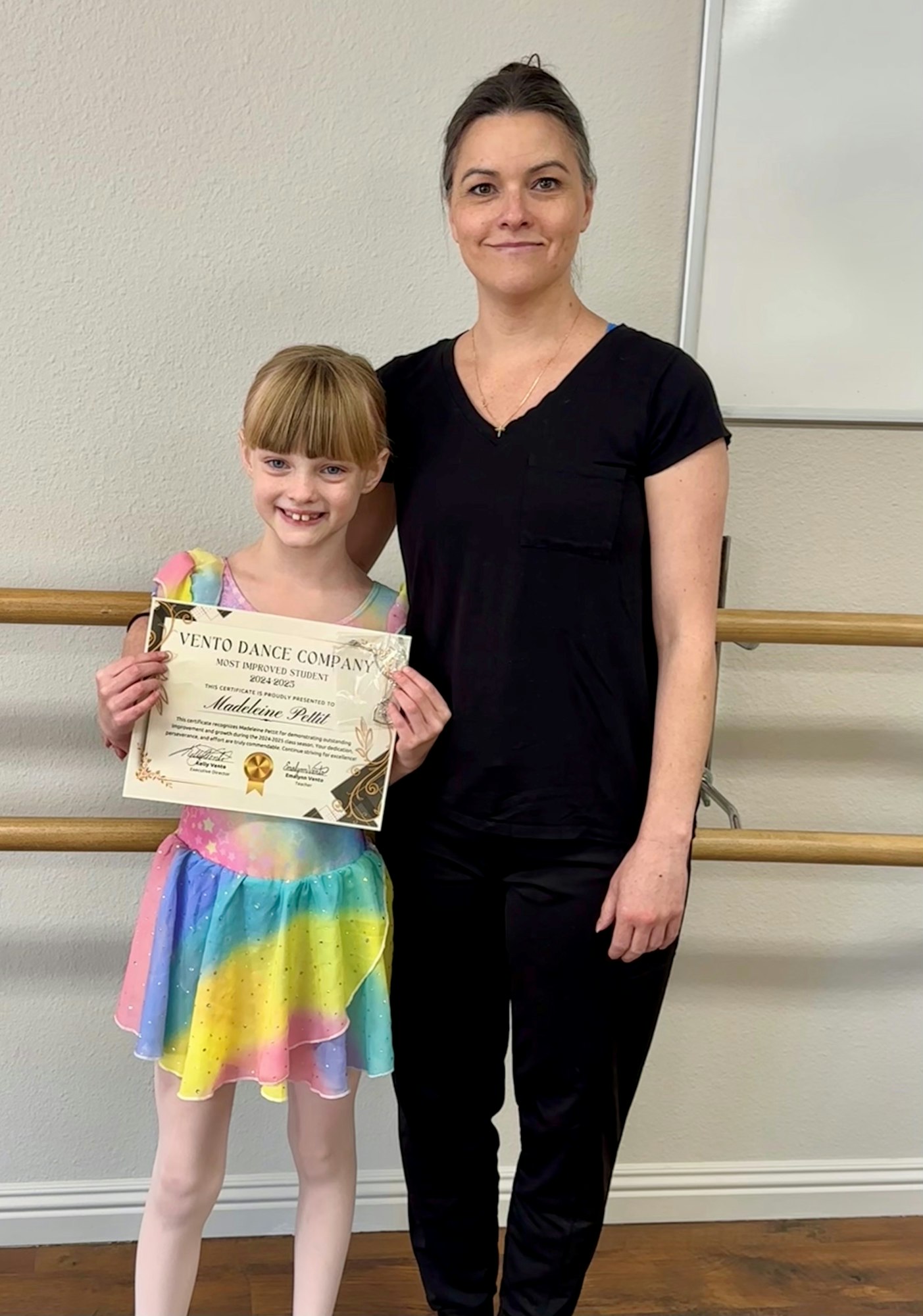 A child in a rainbow dress, holding a dance certificate, stands next to an adult in black against a ballet barre.