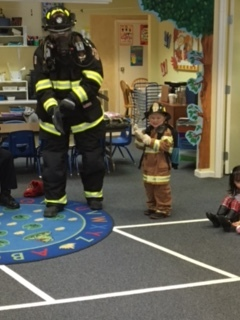 A firefighter in full gear stands with a child dressed in a firefighter costume in a classroom setting.