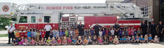 Children and firefighters pose in front of a fire truck labeled "Homer Fire."