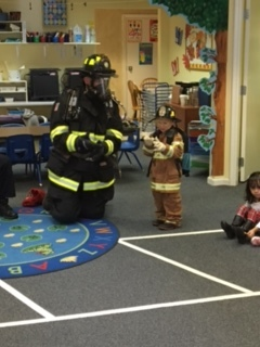 A firefighter and a child dressed in firefighter gear are in a classroom with other kids sitting on the floor.