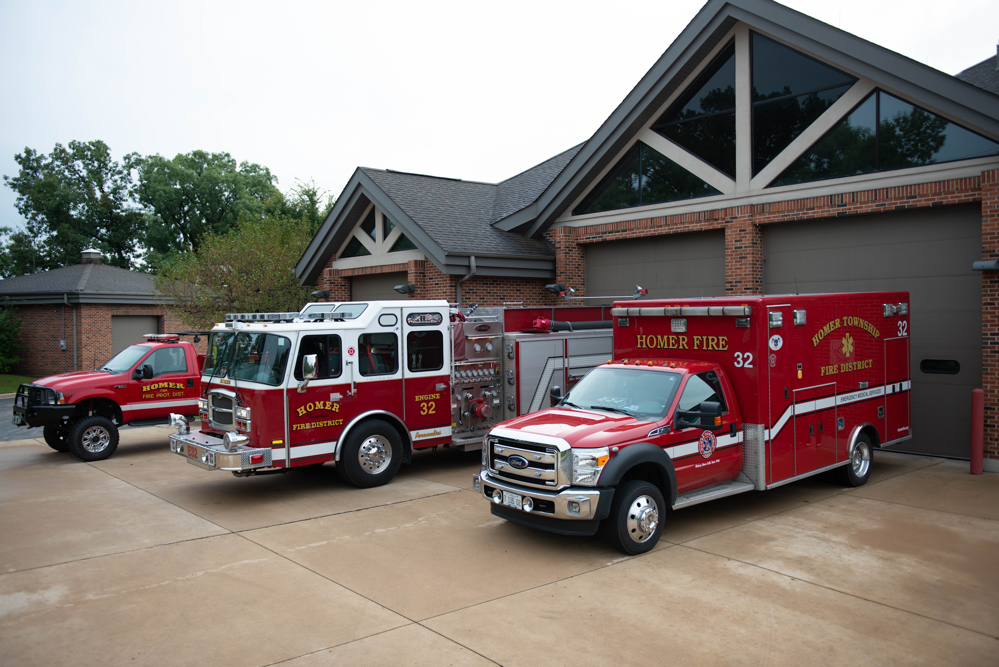 Three red fire vehicles labeled "Homer Fire District" parked outside a fire station.