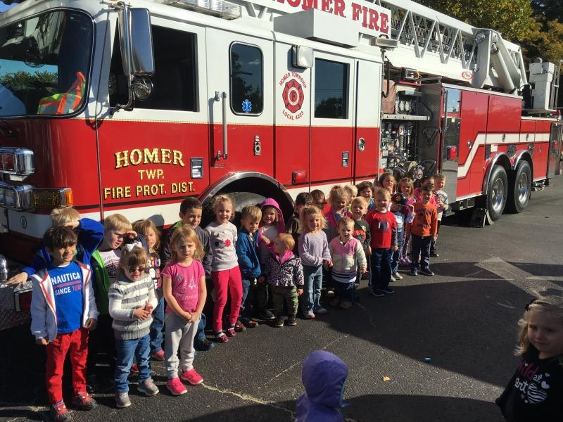 A group of children are standing in front of a red fire truck labeled "Homer Twp. Fire Prot. Dist." on a sunny day.