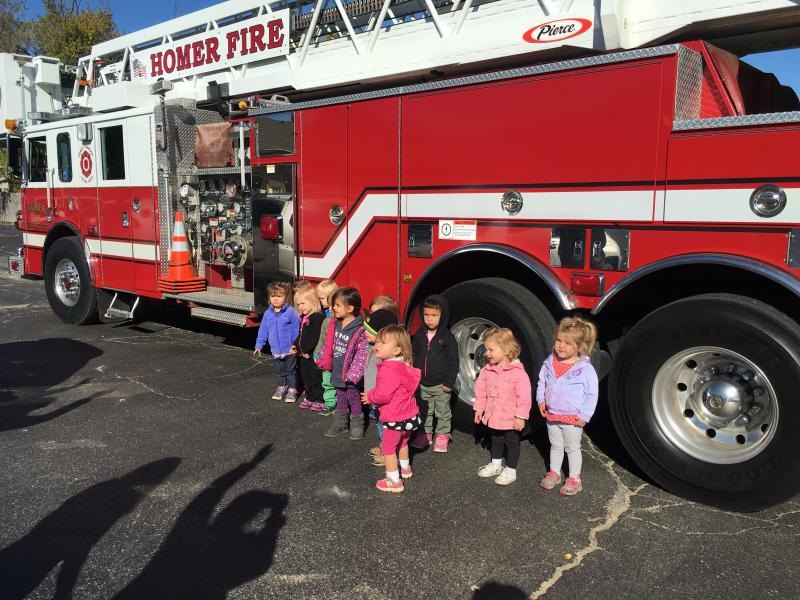A group of children stand in front of a red fire truck marked "Homer Fire" under a sunny sky.