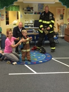 A child dressed as a firefighter is helped by an adult in a classroom with two men, one in a firefighter uniform, observing.