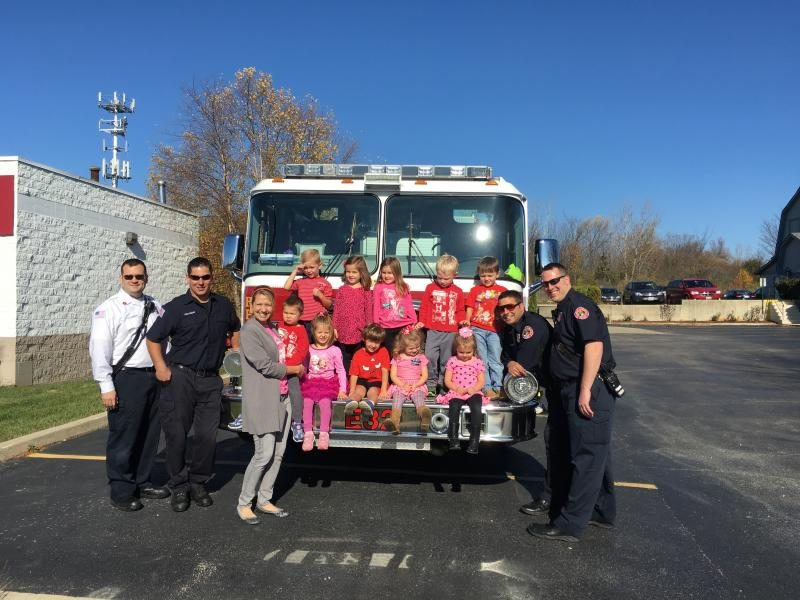 Firefighters and children pose in front of a fire truck in a sunny parking lot.