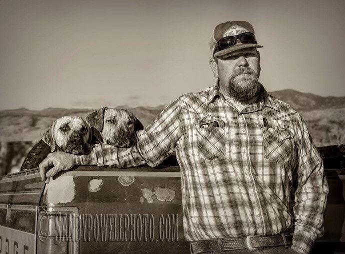 Photo of Jarret Phillips in a baseball cap with his two dogs. Jarret is leaning against the back of his truck.
