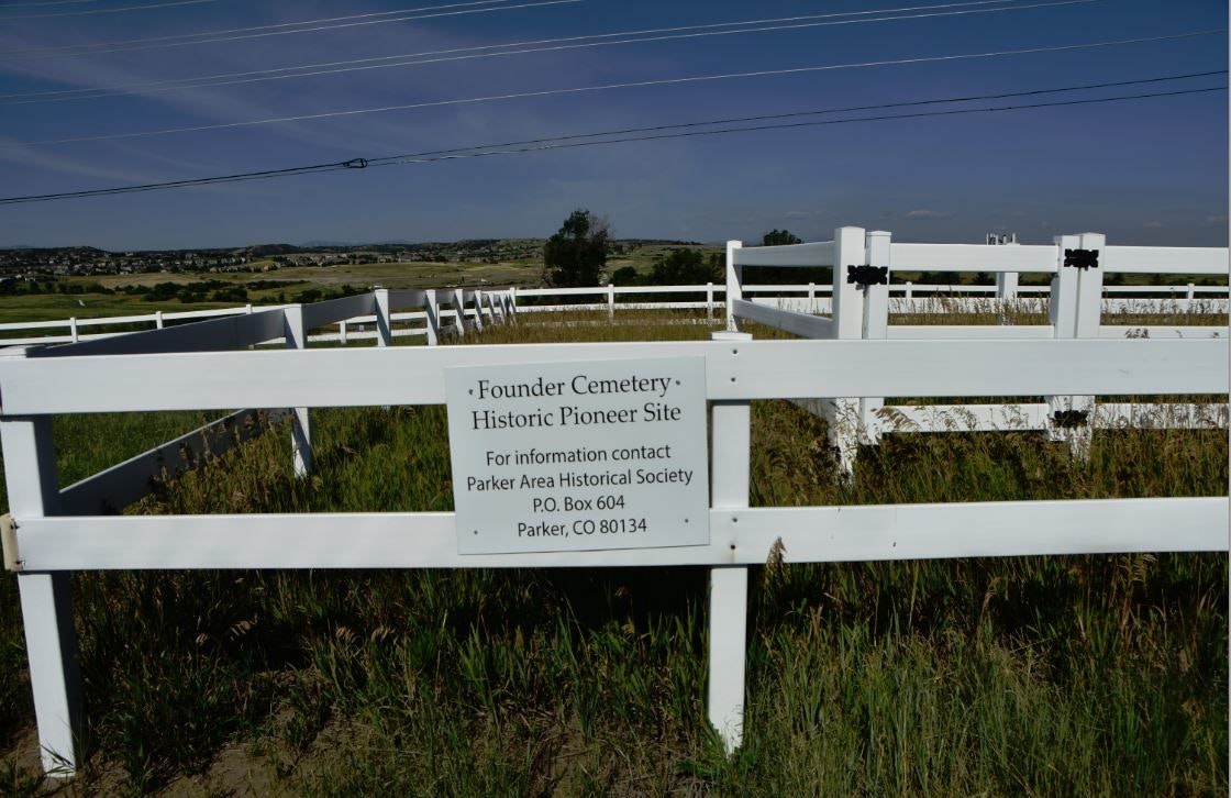 White fenced area with a sign for "Founder Cemetery Historic Pioneer Site" in Parker, CO. Open field and hills in the background.