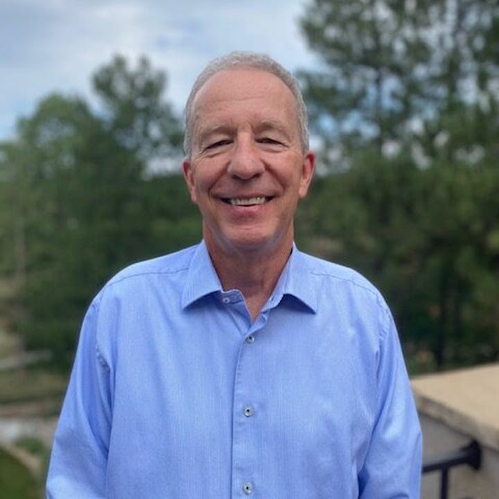 A person in a blue shirt stands outdoors, smiling, with trees and a cloudy sky in the background.